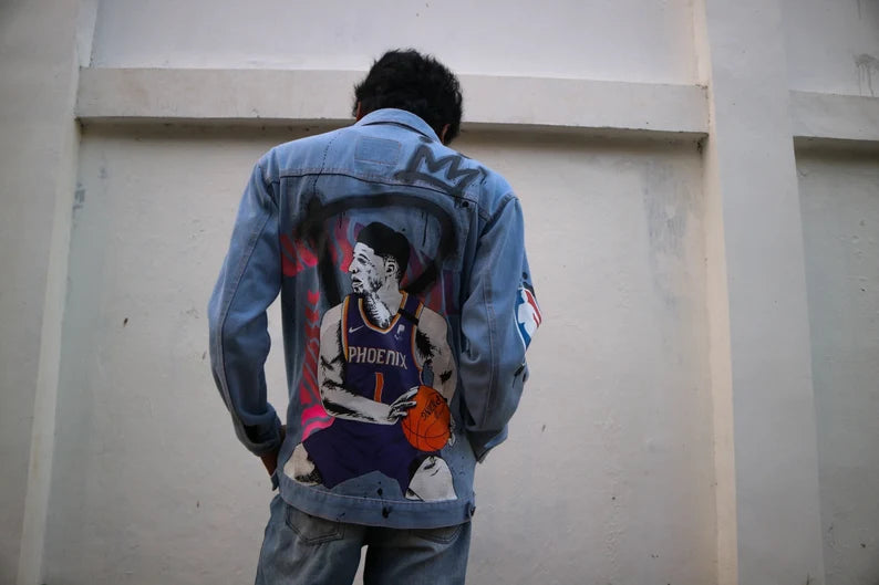 Person wearing a denim jacket featuring a portrait of Devin Booker on the Phoenix Suns with a graphic design on the back against a plain wall.