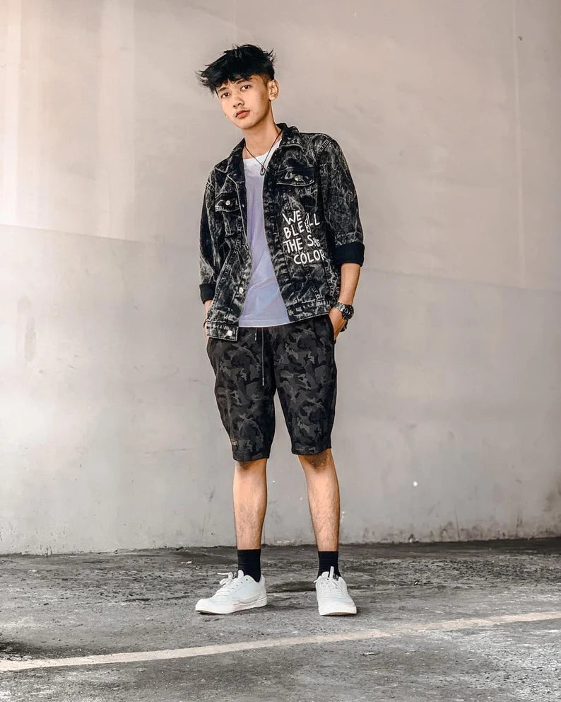 Person wearing a Urban Activist black denim jacket with text, white t-shirt, camouflage shorts, and white sneakers against a plain wall.