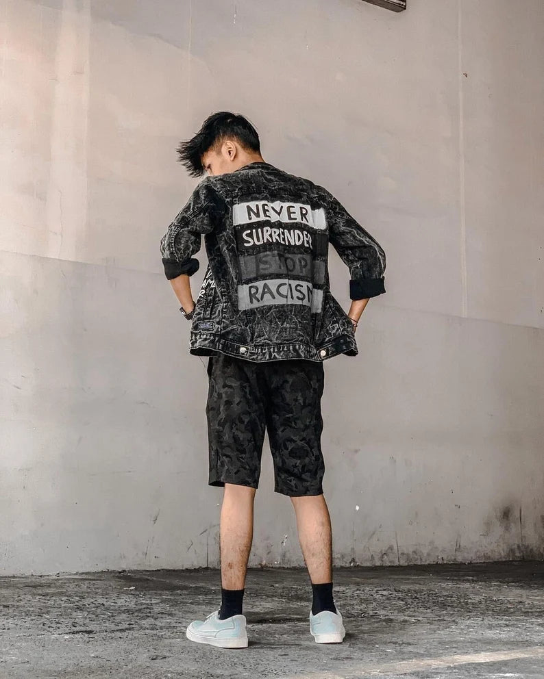 Person wearing a black hand-painted denim jacket with 'Never Surrender' and 'Stop Racism' Social Justice Streetwear