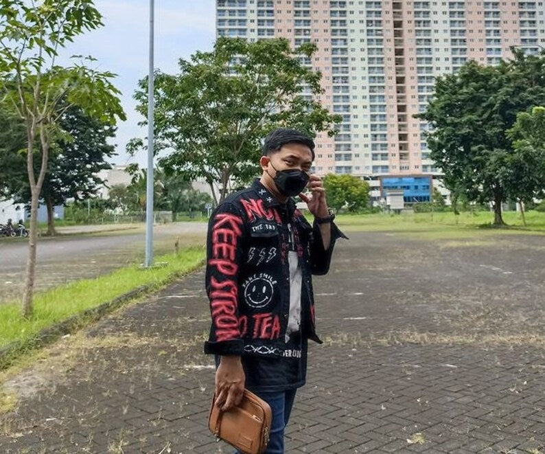 Person wearing a Rock Lover jacket with colorful text and graphics, taking a selfie outdoors with buildings and trees in the background.