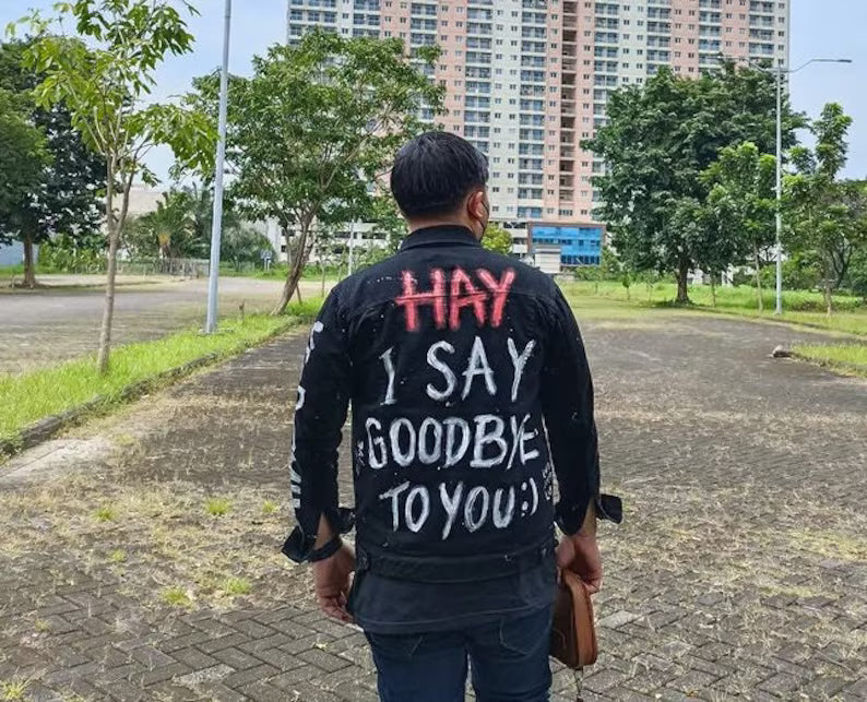 Person wearing a jacket with text on the back, standing in an outdoor setting with buildings and trees in the background.