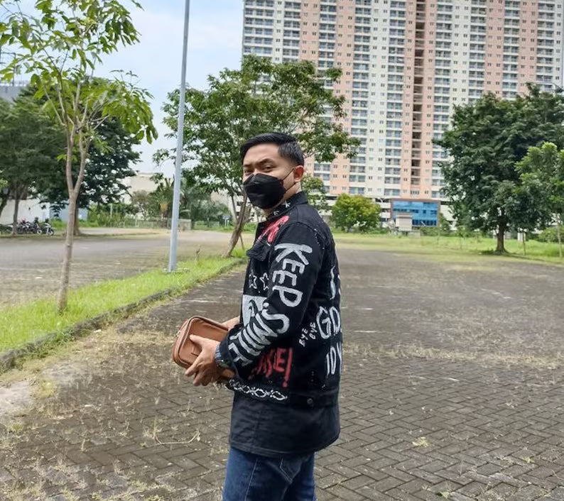 Person wearing a Concert Outfit black jacket with white text and graphics, holding a brown bag, standing on a paved area with trees and buildings in the background.