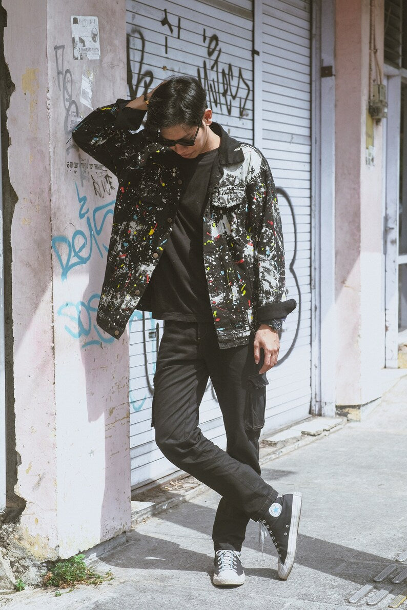 Person wearing a patterned jacket neon paint splatters and black pants standing against a graffiti-covered wall.
