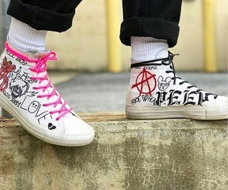 Streetwear White high-top sneakers with black text and designs, worn with pink laces on a concrete surface.