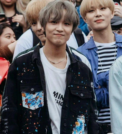Haechan NCT Dream wearing a black jacket with colorful patches and a white shirt, standing in a crowd.