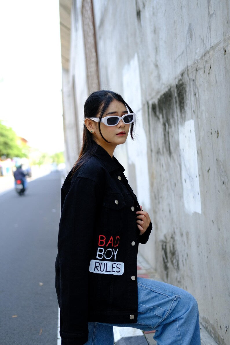 Person wearing a Streetwear black jacket with 'Bad Boy Rules' text, sitting against a concrete wall.