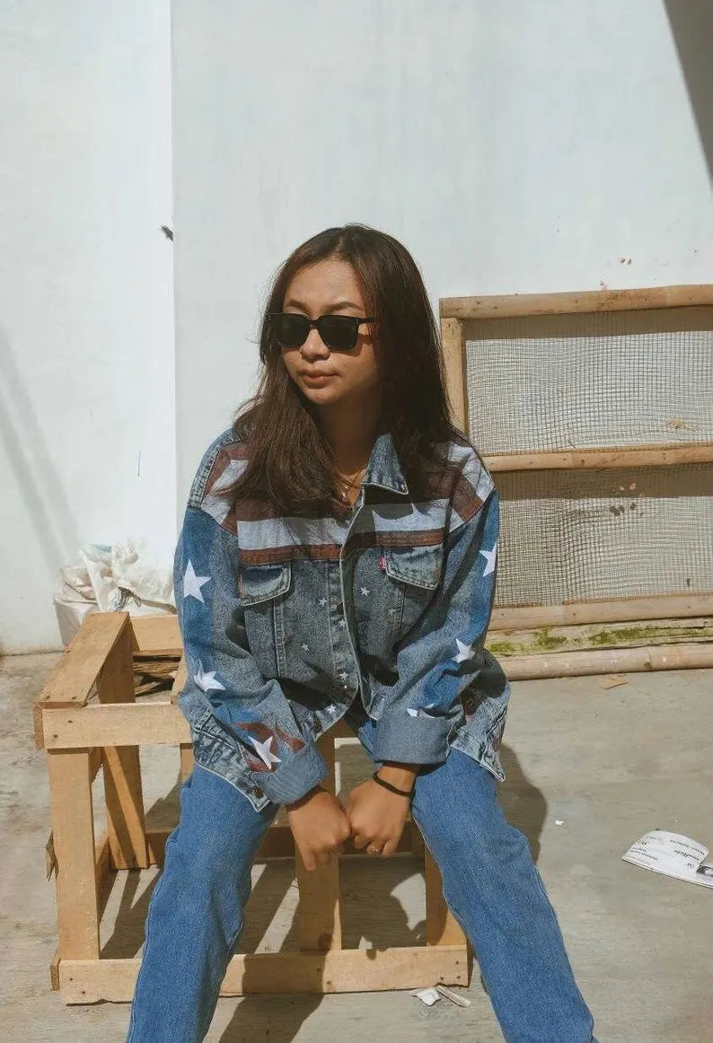 Person wearing a America Chavez Denim Jacket and sunglasses sitting on a wooden crate.